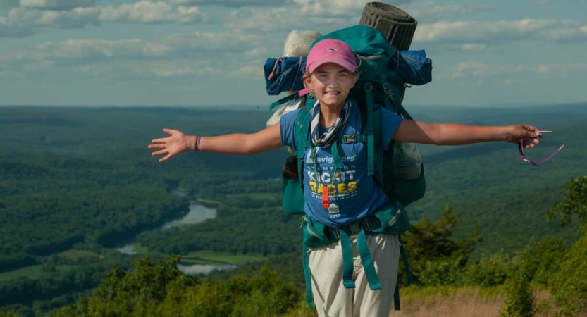 A young person wearing a backpack stands high above a vast green landscape with a river snaking through it. They are holding out their arms wide in apparent celebration.